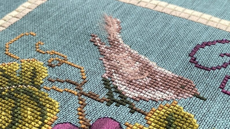 May include: Cross-stitch embroidery depicting a bird with pink and white feathers, perched on a vine with green leaves. The background is a light blue fabric.