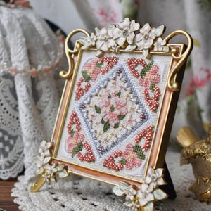 May include: A framed cross-stitch with a floral pattern in pink, red, and white. The gold-coloured frame has flower decorations. The artwork is displayed on a wooden surface. The frame is approximately 15 cm in height.