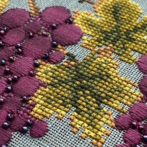May include: Close-up of a cross-stitch embroidery featuring a cluster of purple grapes with dark blue beads. The grapes are surrounded by yellow and green leaves.