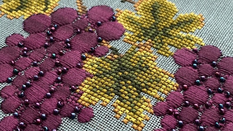 May include: Close-up of a cross-stitch embroidery featuring a cluster of purple grapes with dark blue beads. The grapes are surrounded by yellow and green leaves.
