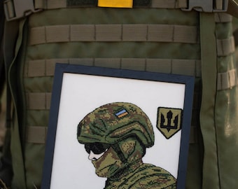 Pode incluir: Um bordado de ponto cruz emoldurado que representa um soldado com um capacete e um uniforme de camuflagem. O soldado está segurando um rifle e tem um remendo da bandeira ucraniana em seu capacete. A moldura é preta e o fundo é branco. O bordado é cercado por uma mochila verde com um remendo da bandeira ucraniana e um remendo de flor amarela.