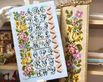 May include: A light blue cross-stitch sampler with the alphabet, floral borders, and a decorative ribbon. A second, vertical floral tapestry with yellow, pink, and purple flowers is also visible. The sampler is held by a gloved hand.