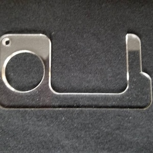 May include: A clear acrylic door opener and button pusher. The tool has a circular opening for a finger, a rectangular section, and a hook-shaped end. The item is designed to avoid direct contact with surfaces.