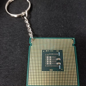 May include: A computer processor keychain. The square-shaped processor has a green base with numerous gold-colored pins. A silver-colored chain and keyring are attached to the processor. The text "004" is visible on the processor.