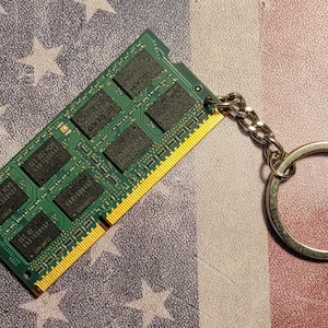 May include: A green RAM memory module keychain with a silver chain and keyring. The RAM module has gold-colored contacts and black chips. The background is a faded American flag.