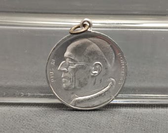 May include: Silver-toned metal pendant featuring a profile portrait of Pope Pius XI. The pendant has a loop at the top for attaching to a chain or cord. The text "PIUS XI" is visible on the left side of the pendant, and "PONTIFEX" is visible on the right side.