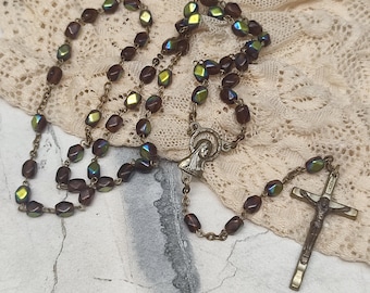 Könnte beinhalten: Ein Vintage-Rosenkranz mit dunkelbraunen, irisierenden Perlen und einem bronzefarbenen Kruzifix. Die Perlen haben eine ovale, facettierte Form. Der Rosenkranz enthält ein Mittelstück mit einer religiösen Figur.