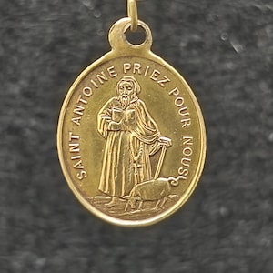 May include: An oval gold-toned metal pendant featuring a relief image of a bearded figure in robes holding a book and a staff, accompanied by a pig. The text SAINT ANTOINE PRIEZ POUR NOUS is embossed along the upper edge of the pendant.