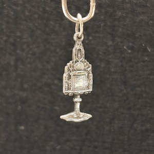 May include: A silver-toned metal charm shaped like a monstrance, featuring an ornate architectural frame with a central circular element, a rectangular base, and a small loop at the top for attachment, suspended from a simple silver-colored metal ring.