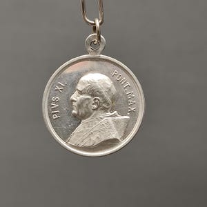 Peut inclure: Médaille ronde argentée avec un portrait de profil. La médaille porte l'inscription "PIVS XI." et "PONT. MAX." sur le bord. Elle est suspendue par une boucle métallique.