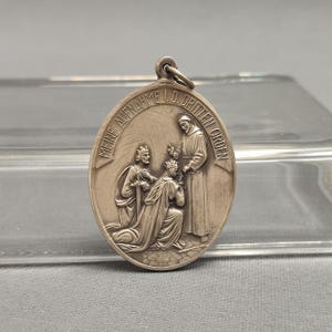 Puede incluir: Pendiente de plata con una escena religiosa que representa a un rey y una reina arrodillados ante un sacerdote. El pendiente tiene forma ovalada y tiene la inscripción "Meine Aufnahme I.D. Dritten Orden" alrededor del borde.