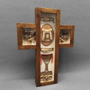 Può includere: Espositore a forma di croce in legno con tre scomparti coperti di vetro. La sezione centrale presenta una figura scolpita e un calice, mentre le sezioni laterali contengono elementi decorativi. La cornice è in legno scuro.