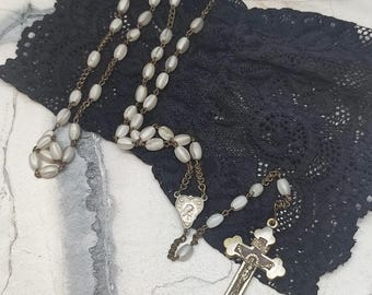 Könnte beinhalten: Ein Vintage-Rosenkranz mit ovalen, cremefarbenen Perlen, die auf einer Bronzekette aufgereiht sind. Der Rosenkranz hat ein Kruzifix und ein herzförmiges Mittelstück. Der Rosenkranz wird auf schwarzem Spitzenstoff präsentiert.