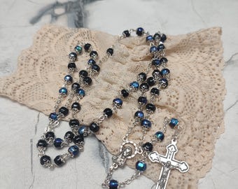 Könnte beinhalten: Eine silberne Rosenkranz mit schwarzen Perlen und einem silbernen Kruzifix. Die Perlen haben eine blaue, irisierende Oberfläche. Der Rosenkranz liegt auf einer weißen Spitzenunterlage.