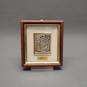 May include: A framed religious plaque with a silver relief depicting a figure with a tree. The frame is dark brown wood with a gold inner border. The word "ASSISI" is on a gold label at the bottom.