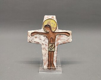 May include: A decorative cross featuring a brown figure with outstretched arms, a yellow halo, and a white and brown cross. The religious symbol is displayed on a clear stand.