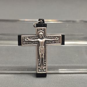 May include: A silver-toned crucifix pendant with a black cross. The figure of Jesus is depicted on the cross, with text inscriptions. The pendant has a small loop for hanging.