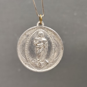 Könnte beinhalten: Ein silberfarbener religiöser Anhänger mit einem detaillierten Bild einer Figur in Gewändern, möglicherweise ein Heiliger. Der Anhänger hat ein ovales Design mit Text an den Rändern und wird von einer Metallkette gehalten.