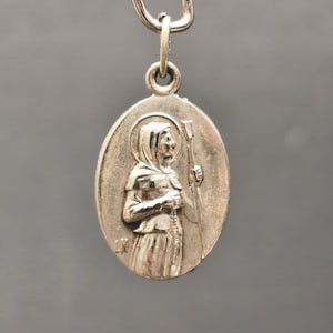 Peut inclure: Pendentif ovale en argent représentant une figure vêtue d'une robe avec un halo, tenant un bâton et un chapelet. Le pendentif est suspendu à une bélière et un fermoir en argent. L'image est sur fond gris.
