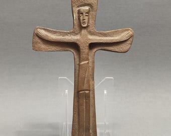 May include: Bronze-colored cross sculpture with a stylized figure. The figure has outstretched arms and is centered on the cross. The sculpture is mounted on a clear acrylic stand against a gray background.