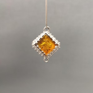 May include: A silver-toned pendant with a diamond-shaped, amber-colored center. The center features a detailed religious emblem. The pendant is suspended from a thin silver wire, and the edges are adorned with a decorative pattern.
