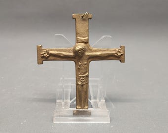 May include: A bronze-colored crucifix pendant featuring a detailed depiction of Jesus Christ. The figure is shown with outstretched arms on the cross. The pendant is displayed on a clear stand against a gray background.