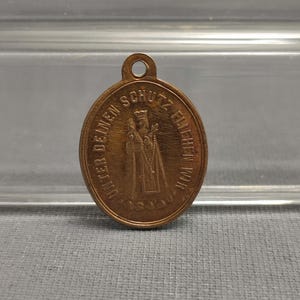 May include: A gold-colored metal pendant with a loop at the top. The front of the pendant features a relief image of a person in a robe holding a staff and a book. The text "Unter deinem Schutz bleiben wir" is inscribed around the image.