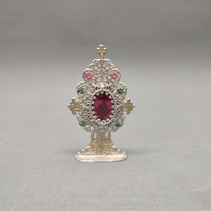 May include: Ornate silver and gold-toned religious reliquary with a large, oval, ruby-colored stone at its center. The reliquary features intricate filigree, pink and green accents, and a small cross at the top. The base is detailed with decorative flourishes.