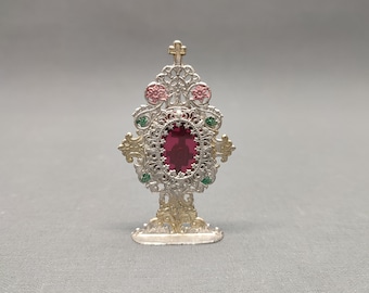 May include: Ornate silver and gold-toned religious reliquary with a large, oval, ruby-colored stone at its center. The reliquary features intricate filigree, pink and green accents, and a small cross at the top. The base is detailed with decorative flourishes.