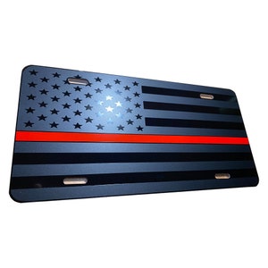 May include: A black and gray American flag with a red stripe across the middle. The flag is on a metal license plate.