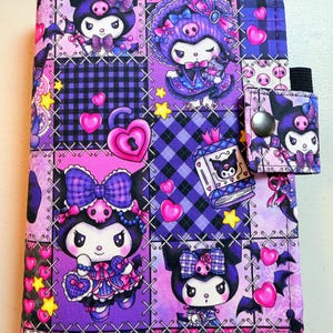 May include: A fabric-covered notebook featuring a repeating patchwork pattern of the character Kuromi in purple, pink, and black. The design includes hearts, stars, and plaid squares, with a snap-button closure tab on the right side featuring Kuromi.