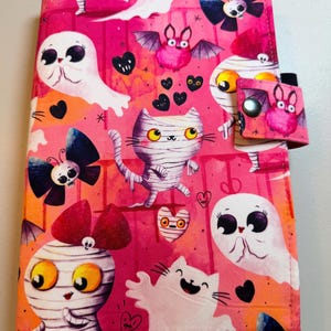 May include: A rectangular fabric book cover featuring a vibrant pink and orange background with a repeating pattern of cartoon ghosts, mummy cats, mummy figures with red bows, bats, and small black hearts, secured by a side tab with a metal snap.
