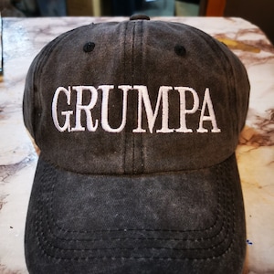May include: A black distressed baseball cap with white embroidered text that reads "GRUMPA".