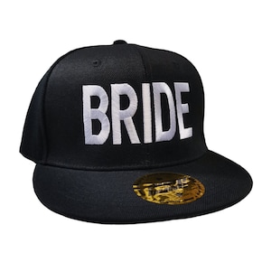 May include: Black baseball cap with the word "BRIDE" embroidered in white. The cap has a flat brim and a gold sticker on the brim. A stylish accessory for a bridal event.