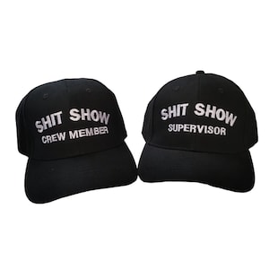 May include: Two black baseball caps with white embroidered text. One cap says "Shit Show Crew Member" and the other says "Shit Show Supervisor".