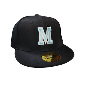 May include: Black flat-brim baseball cap with a large, embroidered letter "M" in light blue and white. The cap has a structured crown and a flat brim. A gold sticker is on the brim.