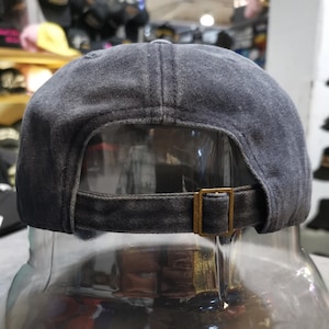 May include: A black denim baseball cap with a brown adjustable strap and buckle. The cap is on a display stand.
