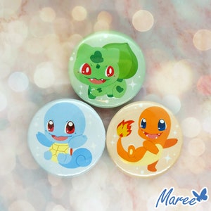 Peut inclure: Trois boutons ronds avec des images de personnages de Pokemon en dessin animé. Les boutons sont vert, bleu et orange. Le bouton vert représente Bulbizarre, le bouton bleu représente Carapuce et le bouton orange représente Salamèche.