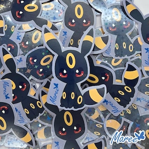 May include: A pile of holographic stickers featuring a black and yellow Pokemon character with a yellow halo. The stickers have a sparkly, iridescent finish and feature the word "Mareee" on some of them.