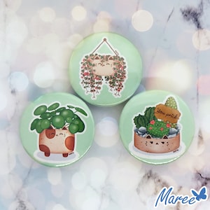 Op de afbeelding: Drie groene en witte ronde knoppen met een kattenpotontwerp. De eerste knop heeft een hangende pot met een kattengezicht en groene wijnstokken. De tweede knop heeft een kattenpot met een groene vetplant. De derde knop heeft een kattenpot met een cactus en de tekst "You prick".