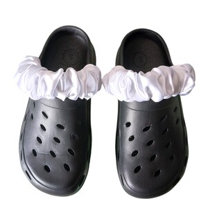 May include: A pair of black clogs with white, ruffled satin fabric accents. The clogs have multiple oval-shaped ventilation holes and a size 5 marking on the inside.