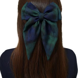 May include: A large fabric bow in a green and blue tartan pattern is attached to the back of a person's head. The bow is a striking accessory against the person's brown hair and dark blue top, creating a bold statement.