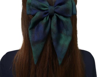 May include: A large, plaid bow hair accessory in shades of green and blue is attached to the back of a person's head. The bow is made of fabric and is a prominent feature against the person's brown hair and dark blue top.