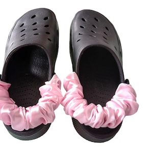 May include: A pair of black clogs with pink satin scrunchie accents. The clogs have multiple ventilation holes and a rounded toe. The scrunchies are a light pink color and are gathered around the front of the clogs.