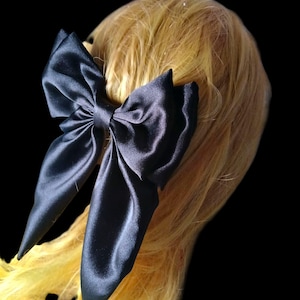 May include: A large, black satin bow hair accessory. The bow is tied and attached to a blonde wig. The bow has long, flowing tails and is positioned on the side of the head. The background is black.