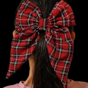 Peut inclure: Un grand nœud rouge à carreaux pour les cheveux. Le nœud est en tissu avec un motif tartan classique rouge, noir et blanc. Le nœud est porté dans les cheveux, le dos du porteur étant tourné vers l'appareil photo, sur fond noir.
