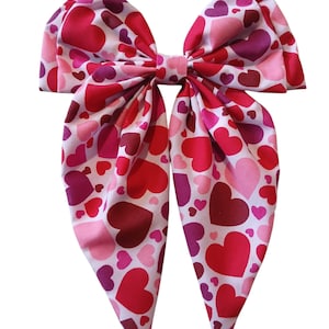 May include: A large pink bow with a heart pattern in shades of pink, red, and burgundy. The bow is made of fabric and has a pointed shape at the ends.