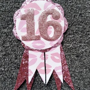 May include: Pink and white glitter ribbon with a pink glitter circle featuring the number 16. The circle has a pink and white lip print pattern.