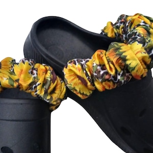 May include: Black clogs with sunflower and leopard print fabric straps. The straps feature yellow sunflowers, green leaves, and a leopard print pattern. The clogs are made of a durable material.
