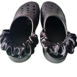 May include: Two black clogs, each featuring a satin ruffle trim around the opening. The clogs have ventilation holes and a ridged sole. The satin ruffles provide a stylish contrast to the casual design of the footwear.